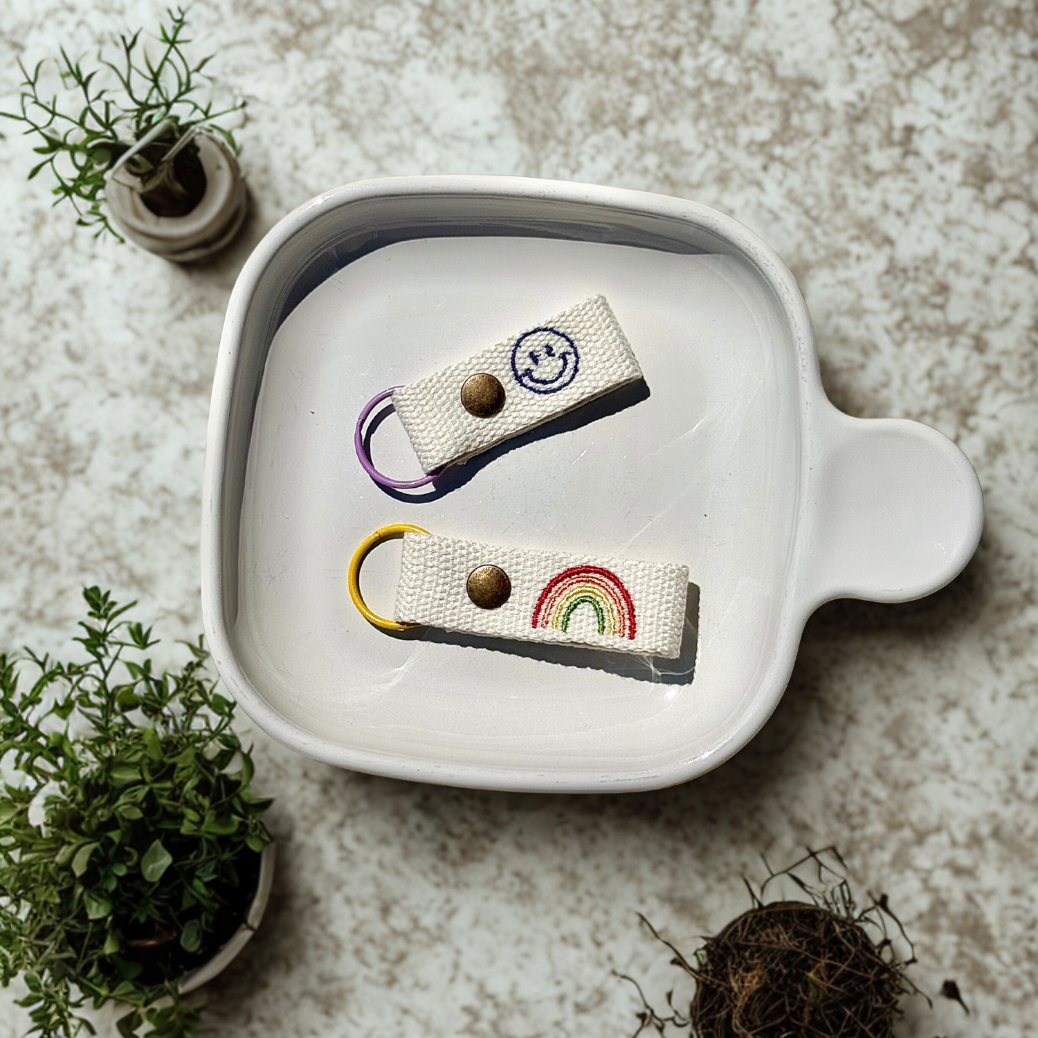 Close-up of handmade fabric keychains featuring a smiley face and rainbow, displayed in a white ceramic dish surrounded by small potted plants. Ideal for showcasing artisanal accessories and DIY crafts.