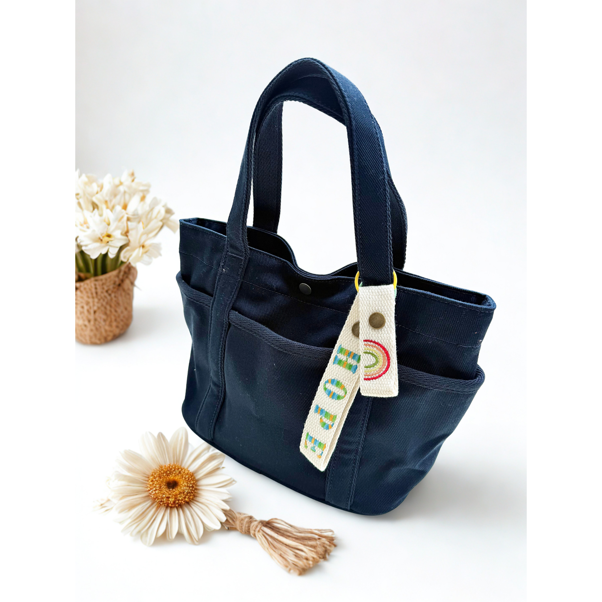 Close-up of a white fabric tag embroidered with ‘HOPE’ in colorful thread and a rainbow icon, attached to a navy blue tote bag—designed by Whimsy Handmade Studio to add personalization and positivity to handcrafted accessories.