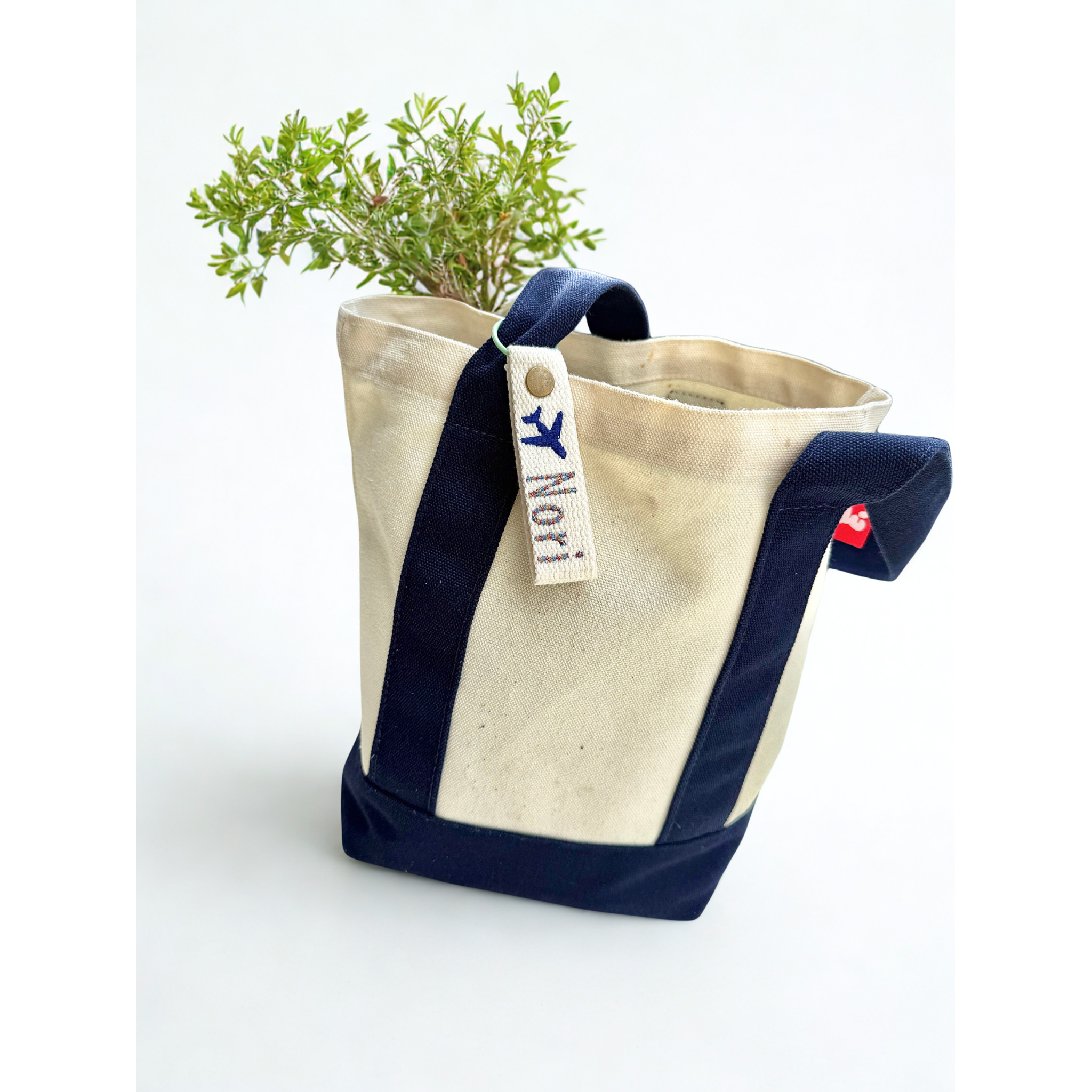 Whimsy Handmade Studio canvas tote bag with black trim and handles, featuring a personalized embroidered tag showing a blue airplane and the name ‘Nori’—a charming detail that highlights artisanal stitching and thoughtful customization
