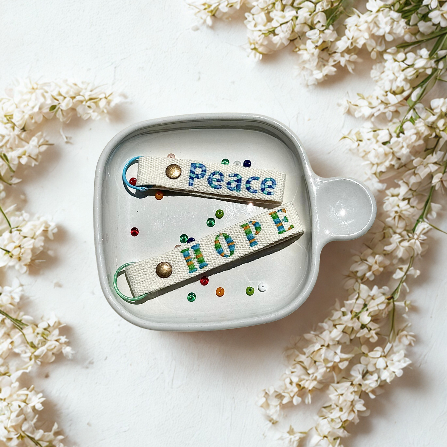 Whimsy Handmade Studio embroidered keychains reading ‘Peace’ and ‘Hope’ nestled in a white dish, surrounded by delicate white flowers and colorful beads—symbolizing serenity and optimism through artisanal craftsmanship