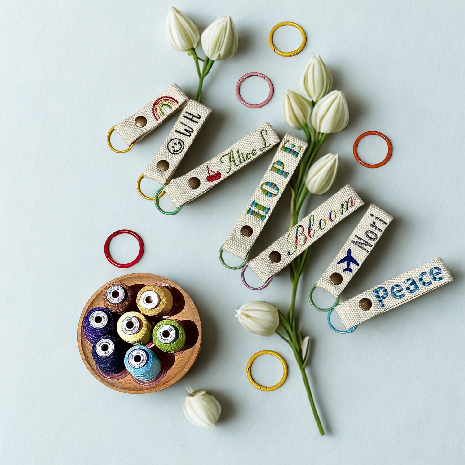 Whimsy Handmade Studio embroidered keychains featuring words like ‘Alice L,’ ‘Hope,’ ‘Bloom,’ ‘Nori,’ and ‘Peace’ displayed on a light background, surrounded by white flowers, colorful rings, and a wooden bowl filled with vibrant thread spools—capturing the charm and detail of personalized, handcrafted accessories.