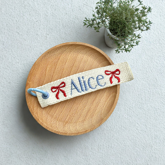 Custom embroidered name tag with “Alice” in blue thread and red bows on white fabric — perfect for personalized clothing labels, kids’ accessories, and unique gift ideas.